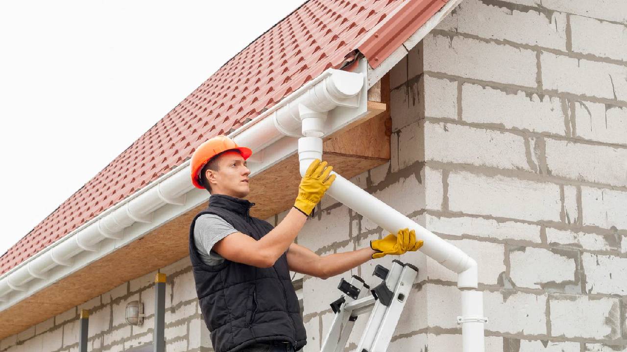 Worker in orange hard hat and yellow gloves installing white gutters on brick house with red tile roof.