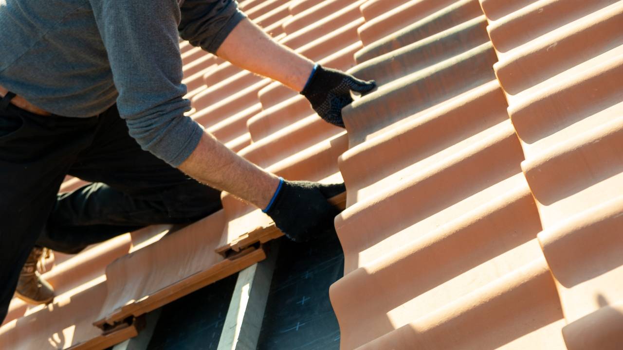 Roofer installing clay roof tiles, kneeling on wooden battens while positioning terracotta tiles in overlapping rows.
