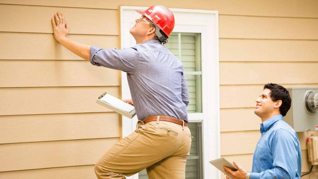 Home inspector in red hard hat examining exterior siding while homeowner with tablet watches from below.