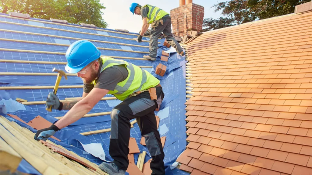 Two roofers in blue hard hats and safety vests installing clay tiles over blue underlayment on residential roof.