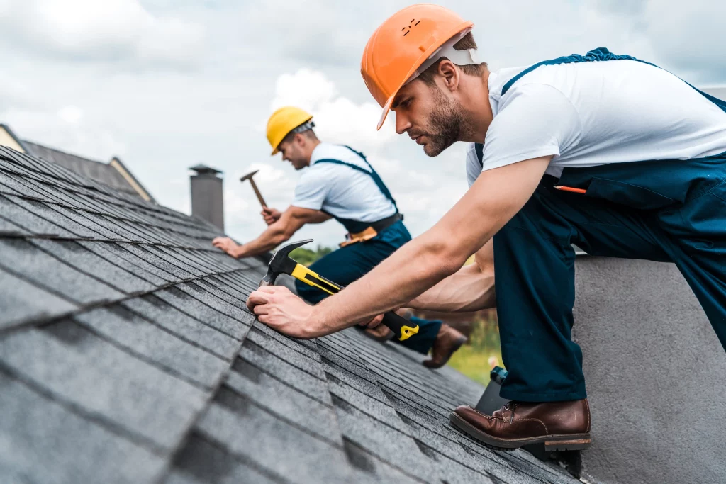 Two roofers in hard hats working on asphalt shingles - one using power tool in foreground, another with hammer behind.